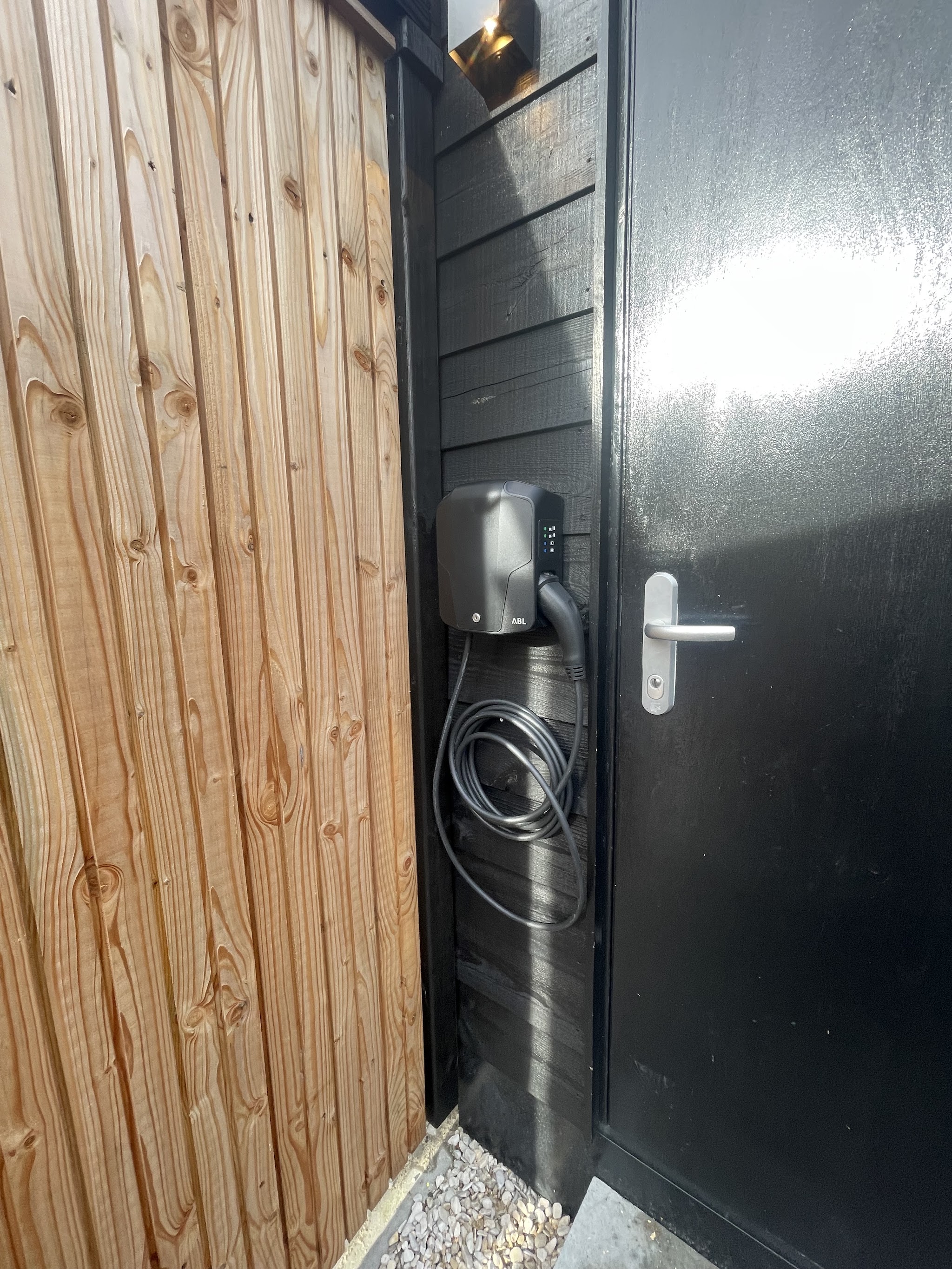 Laadpaal aan de gevel naast een zwarte deur en een houten schutting
